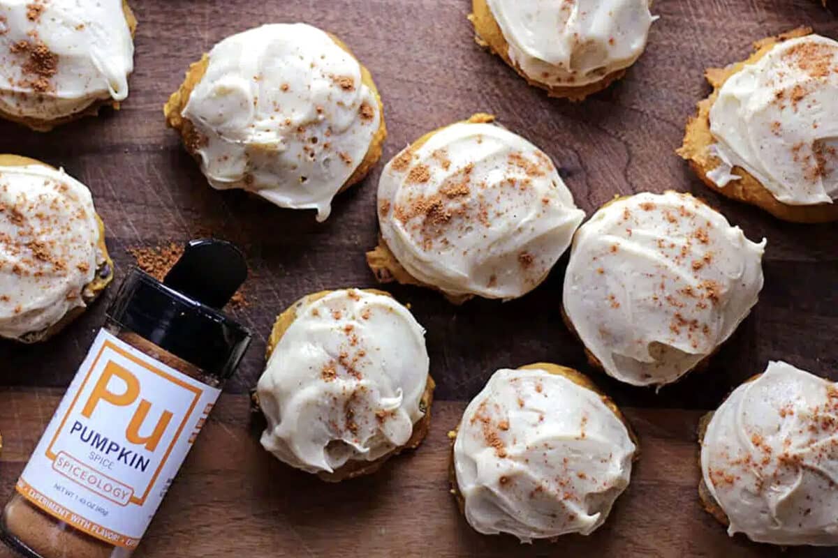 Pumpkin spiced cookies topped with white frosting and a sprinkle of spice are arranged on a wooden surface. A container labeled Pumpkin Spiceology lies nearby, partially open, with some aromatic spice spilled onto the cookies.