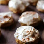 Pumpkin spiced cookies with creamy white frosting and a sprinkle of cinnamon sit on a wooden surface, with several cookies in soft focus in the background.