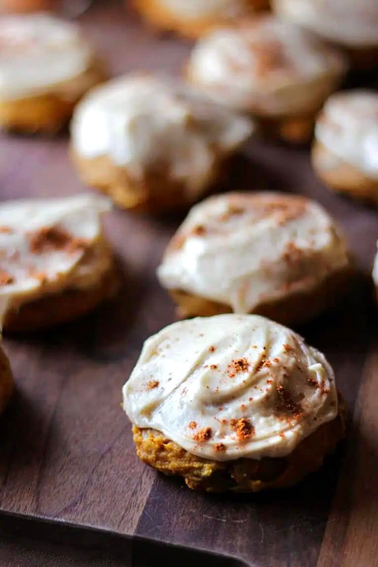 Pumpkin spiced cookies with creamy white frosting and a sprinkle of cinnamon sit on a wooden surface, with several cookies in soft focus in the background.
