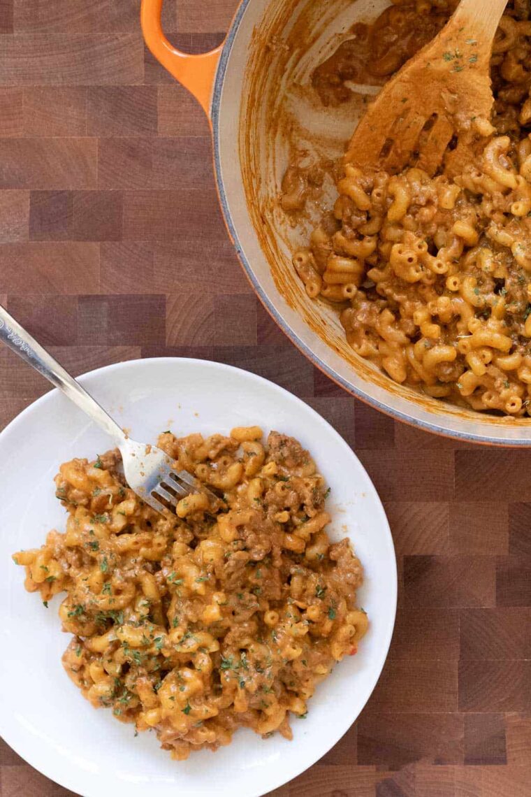 A white plate with a serving of cheesy macaroni and ground beef, resembling homemade hamburger helper, sits next to a pot of the same dish on a wooden surface. A fork rests on the plate, and the food is garnished with herbs.
