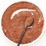 A white bowl filled with coarse, reddish-brown homemade taco seasoning, with a black spoon resting inside and holding some of the spice mixture. The bowl is set on a white surface.