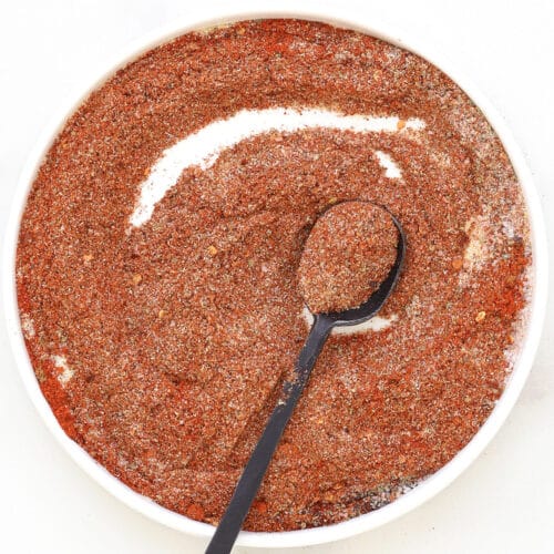 A white bowl filled with coarse, reddish-brown homemade taco seasoning, with a black spoon resting inside and holding some of the spice mixture. The bowl is set on a white surface.
