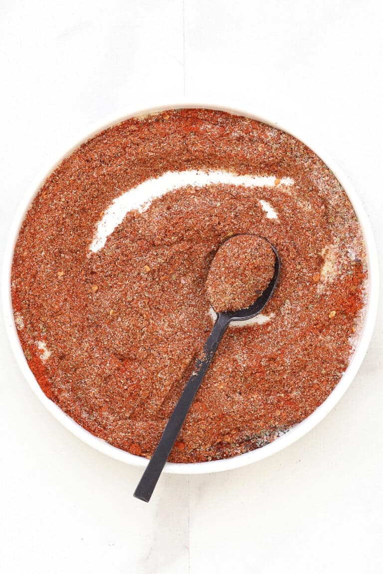 A white bowl filled with coarse, reddish-brown homemade taco seasoning, with a black spoon resting inside and holding some of the spice mixture. The bowl is set on a white surface.