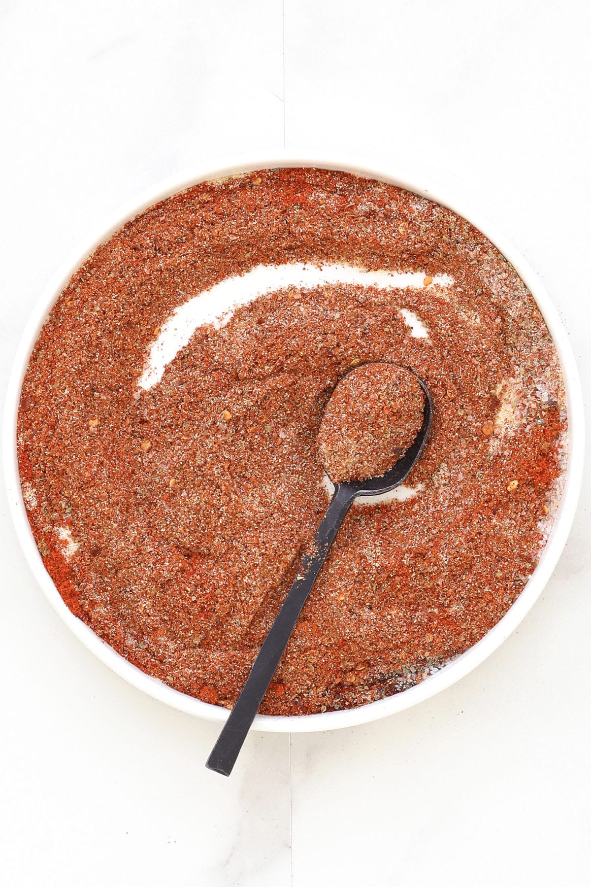 A white bowl filled with coarse, reddish-brown homemade taco seasoning, with a black spoon resting inside and holding some of the spice mixture. The bowl is set on a white surface.