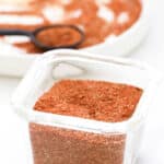 A small glass jar filled with a reddish-brown homemade taco seasoning sits on a white surface, with a white plate and a measuring spoon containing more of the spice mix in the background.