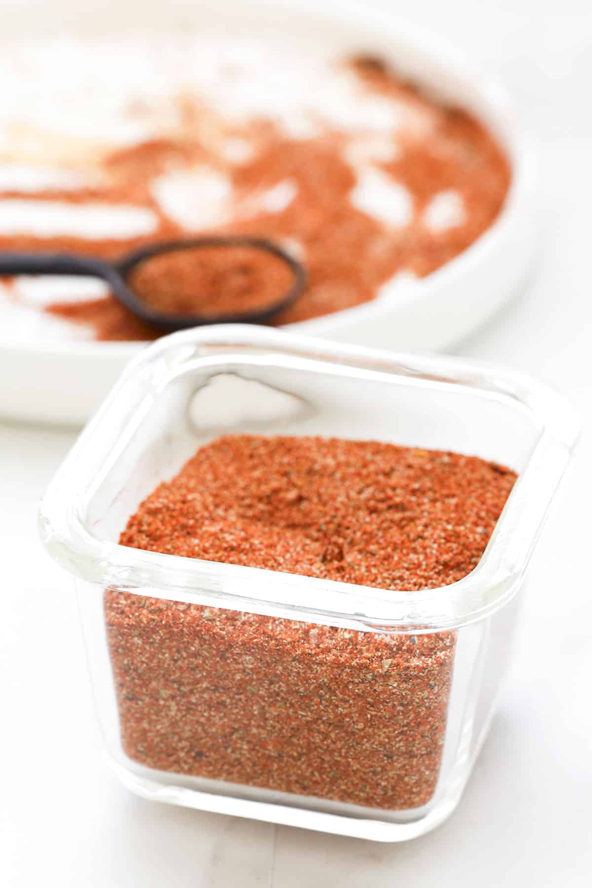 A small glass jar filled with a reddish-brown homemade taco seasoning sits on a white surface, with a white plate and a measuring spoon containing more of the spice mix in the background.