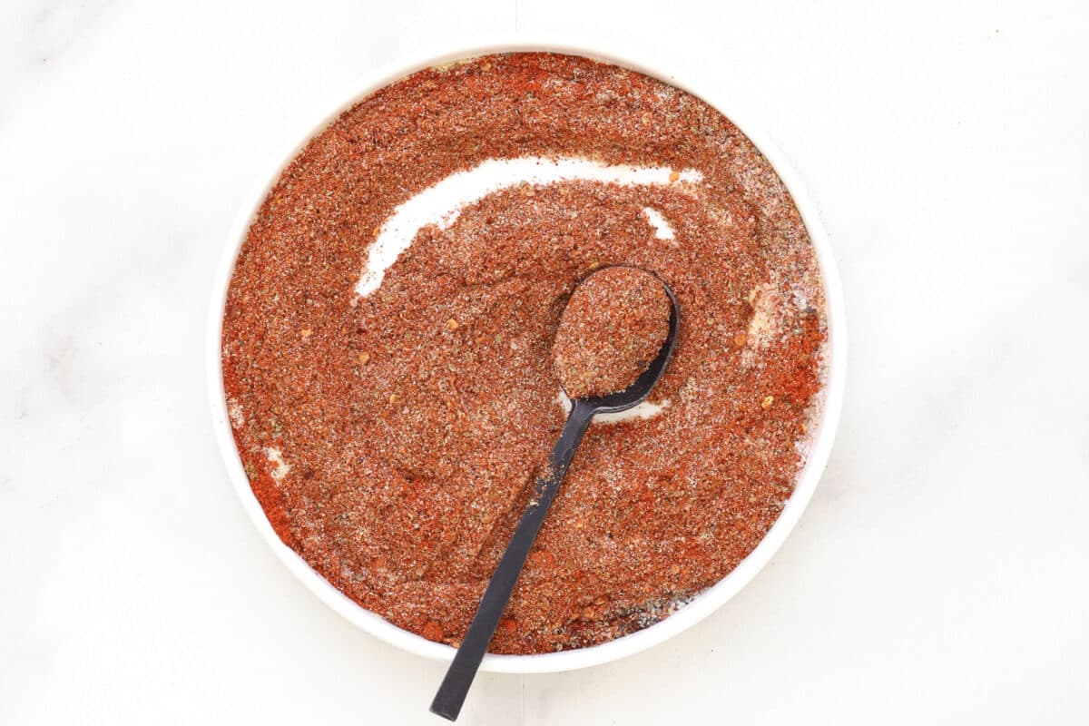 A white bowl filled with homemade taco seasoning, its reddish-brown blend scooped up by a black spoon resting inside. The bowl sits on a clean white surface.