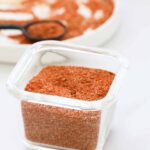 A clear glass container filled with homemade taco seasoning sits on a white surface, with a plate and a black measuring spoon holding more of the vibrant reddish-brown spice blend in the blurred background.