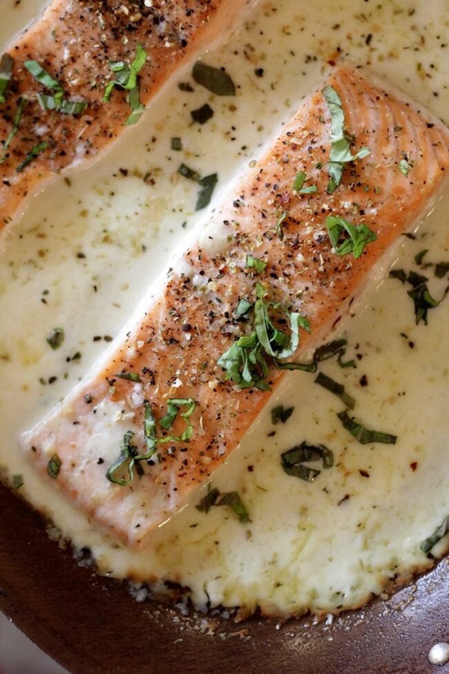 Two baked salmon fillets cooked in a creamy white sauce, topped with cracked black pepper and chopped fresh herbs, in a skillet viewed from above.