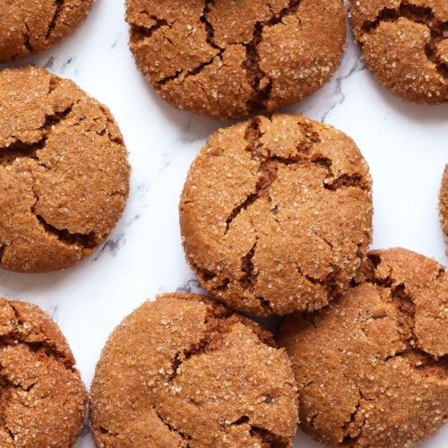 A close-up of several round, cracked ginger cookies with fresh ginger, coated in sugar and arranged on a white marble surface.