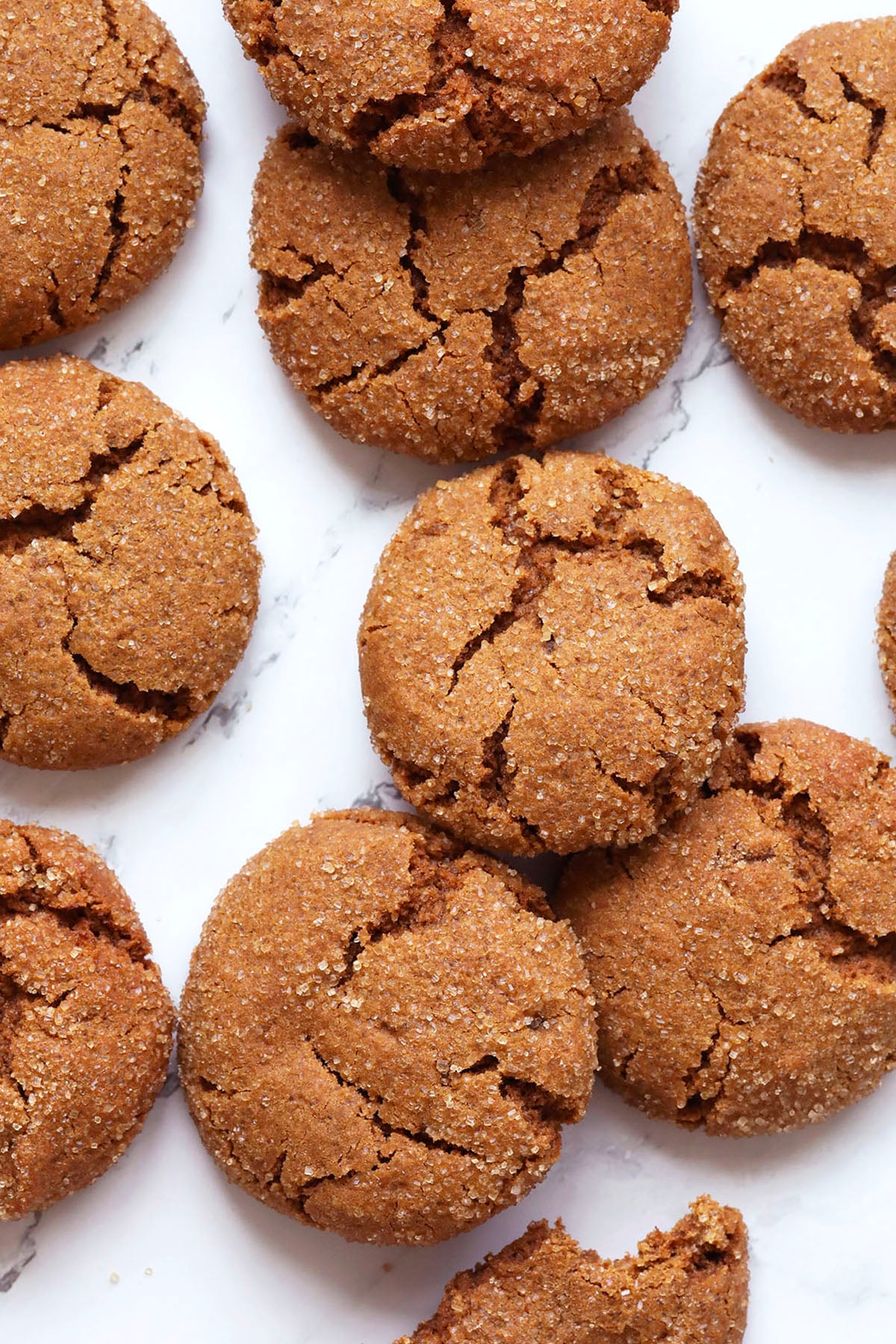A close-up of several round, cracked ginger cookies with fresh ginger, coated in sugar and arranged on a white marble surface.