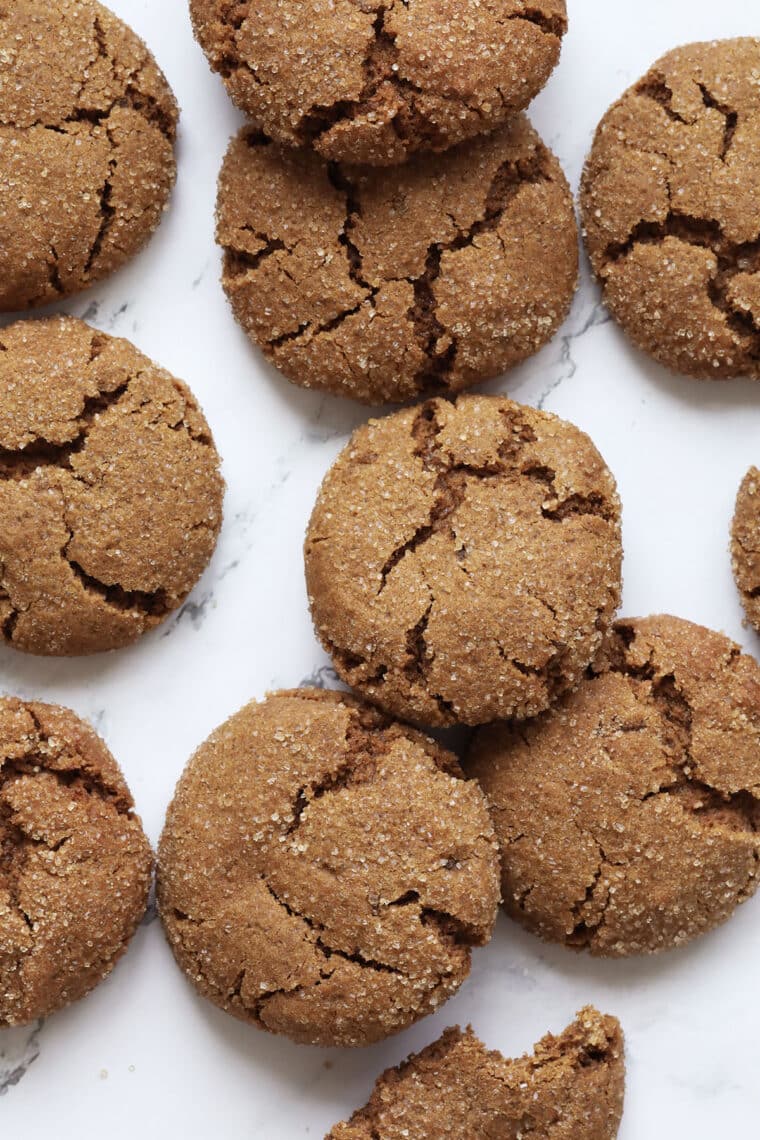 Close-up of several round, golden-brown ginger cookies with crackled tops, arranged on a white marble surface. The cookies have a sugar-coated appearance and rough texture.