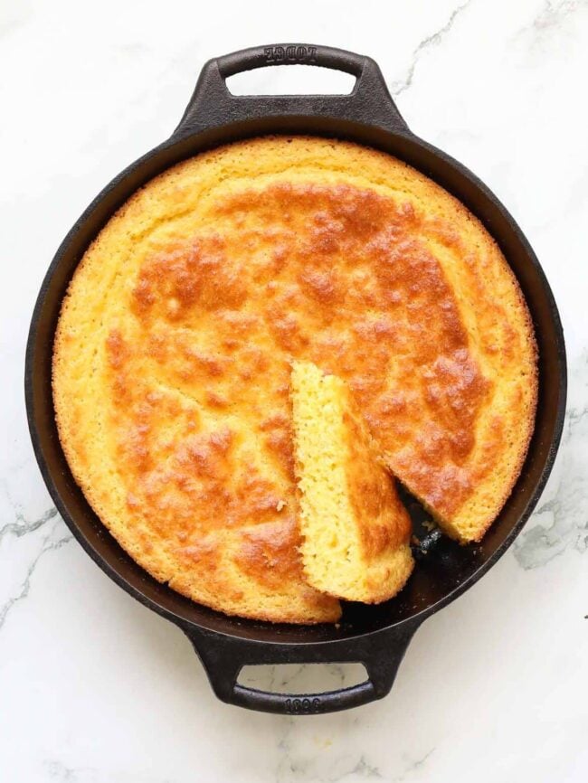 A golden-brown buttermilk cornbread baked in a black cast iron skillet, with one triangular slice partially removed, sits on a white marble surface.