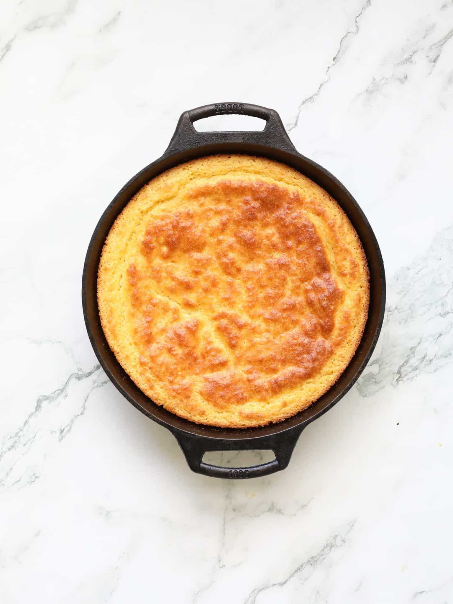A round, golden-brown buttermilk cornbread sits in a black cast iron skillet on a white marble surface, viewed from above.