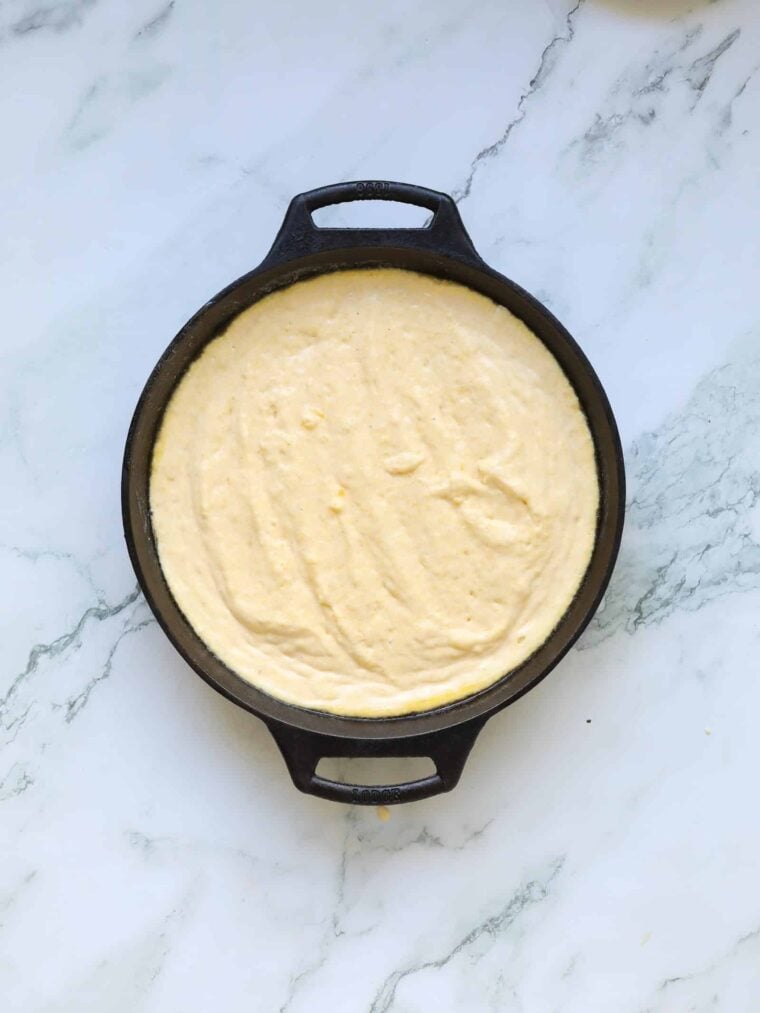 A round cast iron skillet filled with uncooked buttermilk cornbread batter sits on a white marble surface.