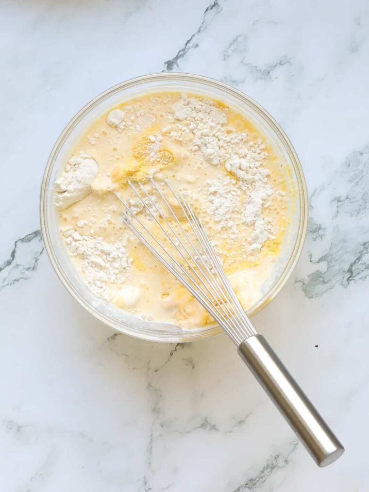 A glass bowl filled with flour, eggs, and milk mixture for buttermilk cornbread sits on a marble countertop; a metal whisk rests inside, ready for mixing.
