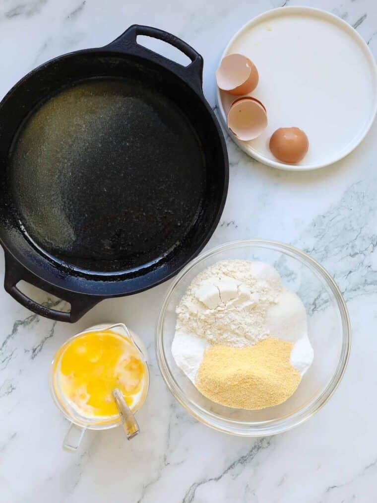 A greased cast iron skillet, two cracked eggshells on a white plate, a glass bowl with flour, cornmeal, sugar, and baking powder for buttermilk cornbread, plus a measuring cup with beaten eggs and milk, all on a marble surface.