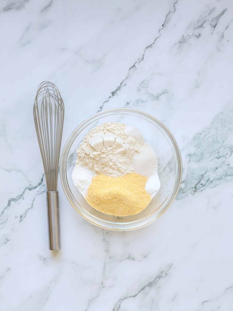 A metal whisk lies next to a glass bowl holding separated piles of ingredients—flour, cornstarch, and cornmeal—on a white marble surface, ready for classic buttermilk cornbread.