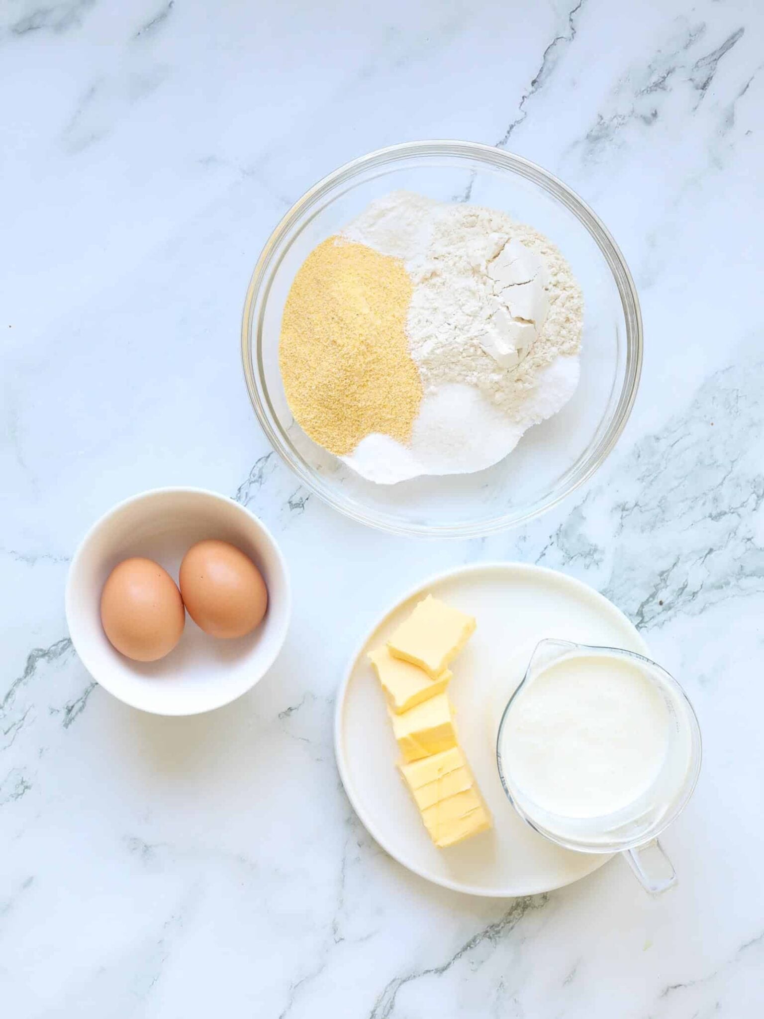A marble countertop displays the ingredients for buttermilk cornbread: one bowl with flour, cornmeal, and sugar; another with two eggs; plus a plate of sliced butter and a small pitcher of milk.