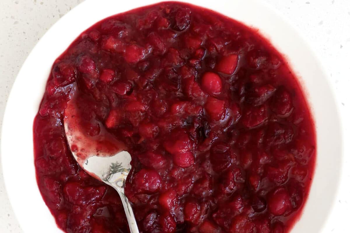 A white bowl filled with chunky red cranberry sauce, with a silver spoon resting inside the bowl, showcases the fresh and vibrant appeal of homemade cranberry sauce.