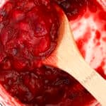 A wooden spoon stirs a pot of chunky red cranberry sauce, with vibrant cranberry sauce smeared on the white sides of the pot.