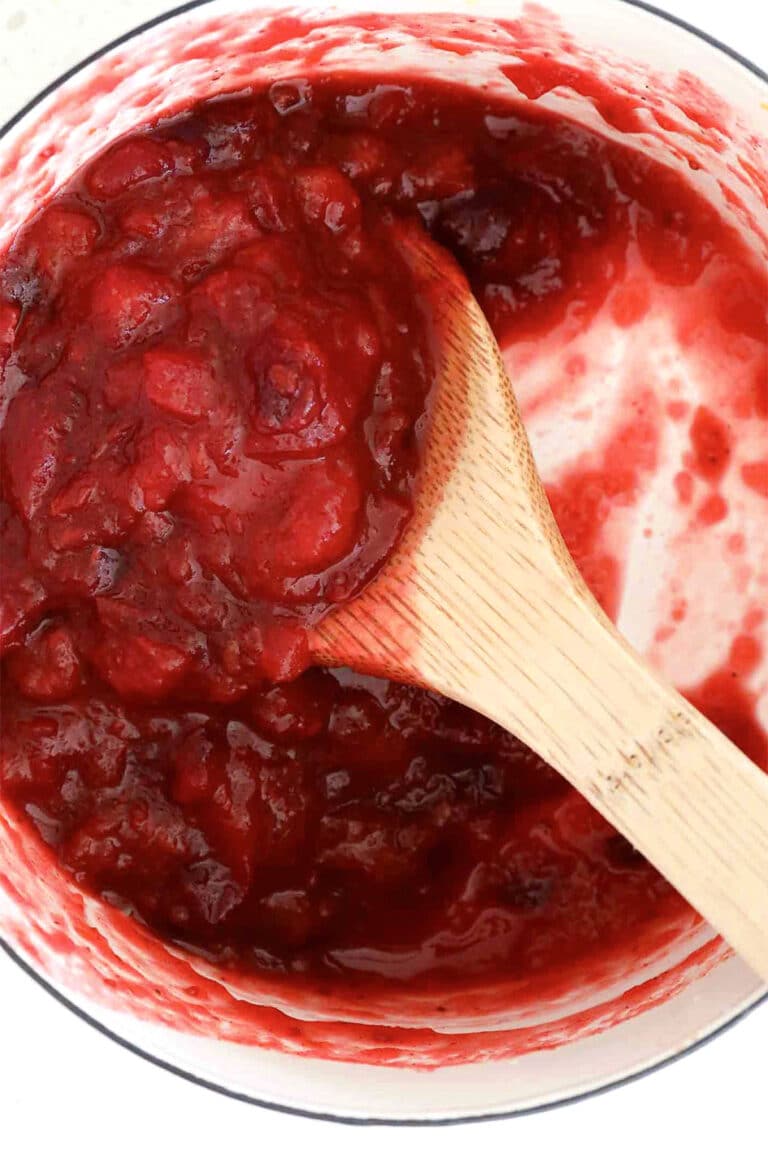 A wooden spoon stirs a pot of chunky red cranberry sauce, with vibrant cranberry sauce smeared on the white sides of the pot.