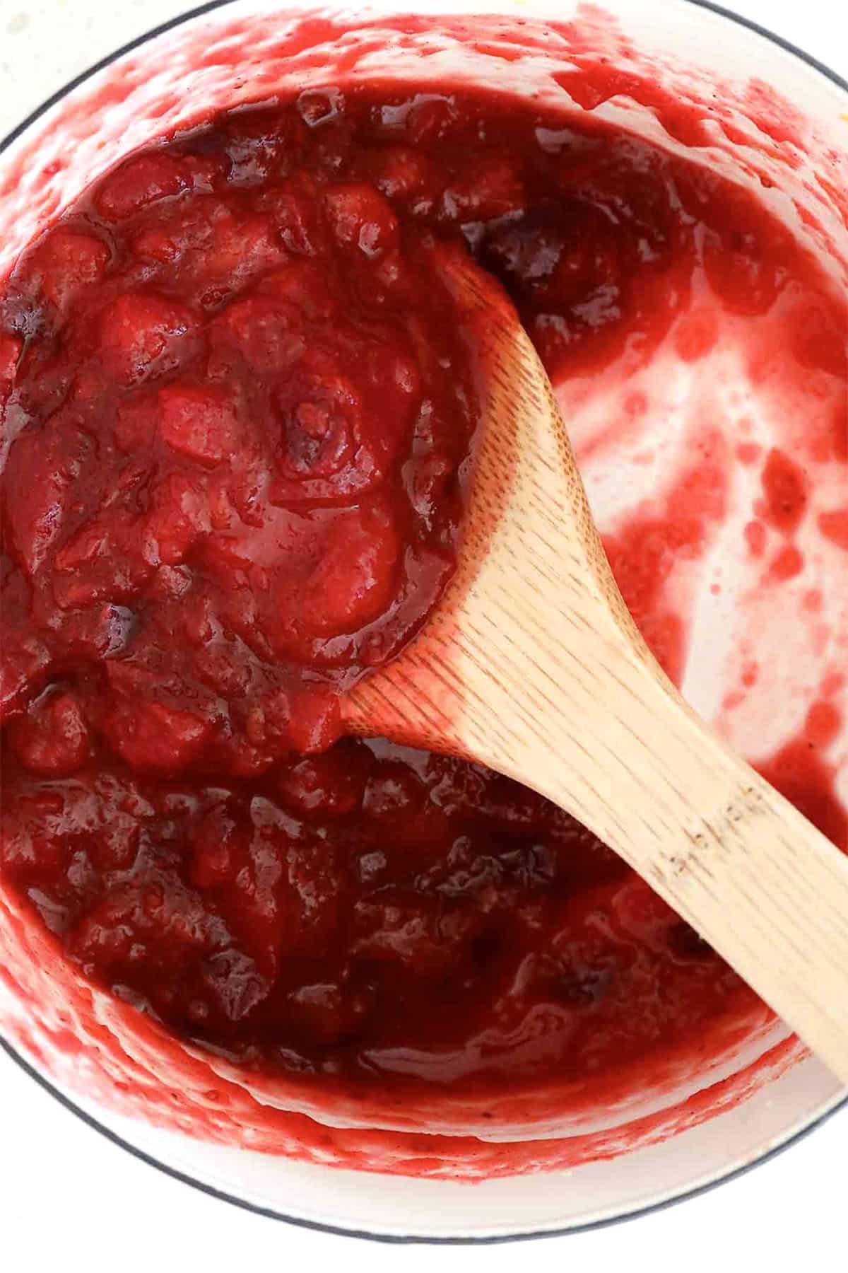A wooden spoon stirs a pot of chunky red cranberry sauce, with vibrant cranberry sauce smeared on the white sides of the pot.