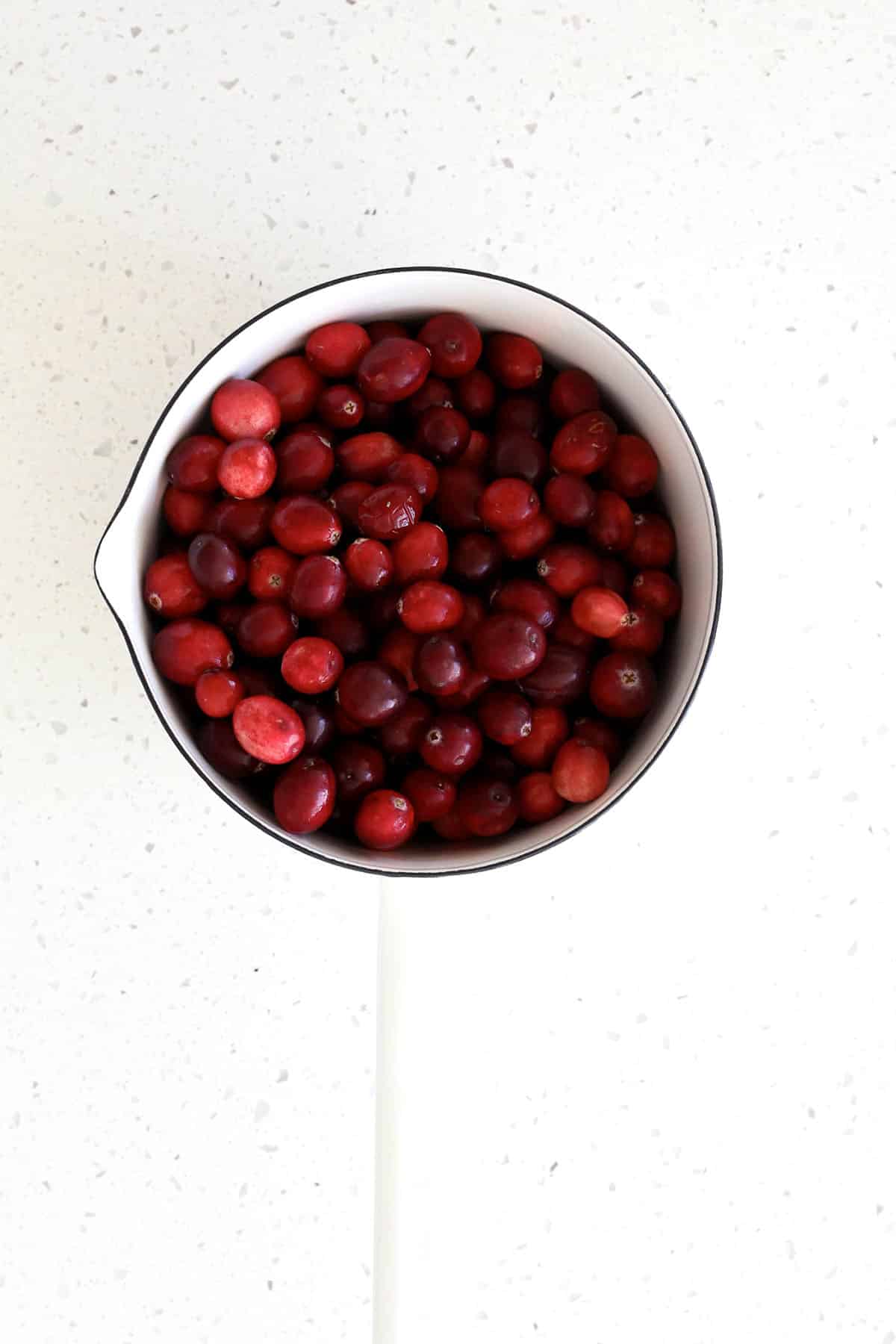 A white saucepan filled with fresh red cranberries, perfect for making homemade cranberry sauce, sits on a white speckled countertop.
