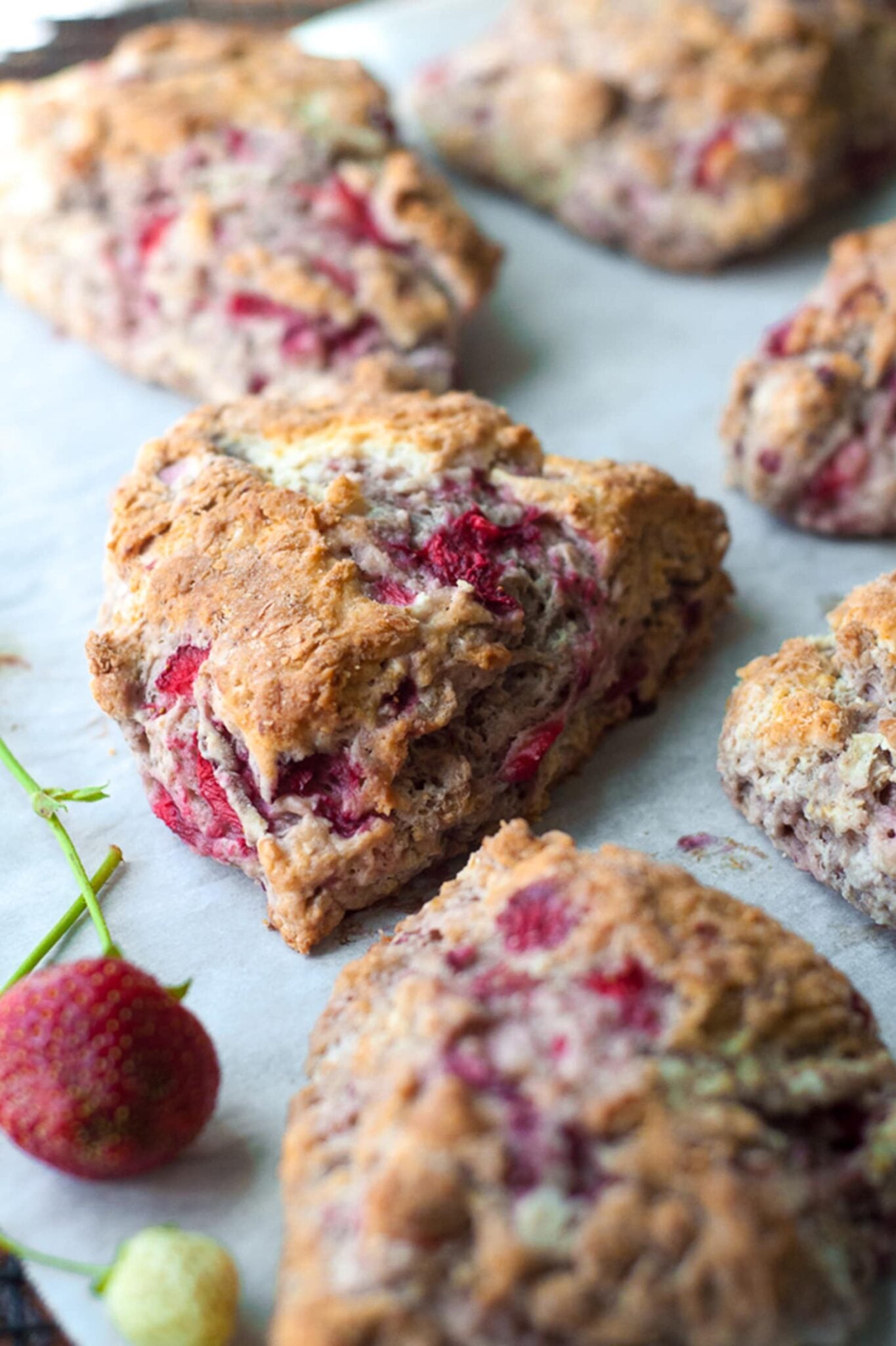 Close-up of fresh strawberry scones with visible pieces of red strawberries, placed on parchment paper. Whole strawberries are scattered around the scones.