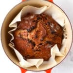 A round loaf of cinnamon raisin bread with chocolate chips sits on parchment paper inside an orange Dutch oven, viewed from above on a white marble surface.