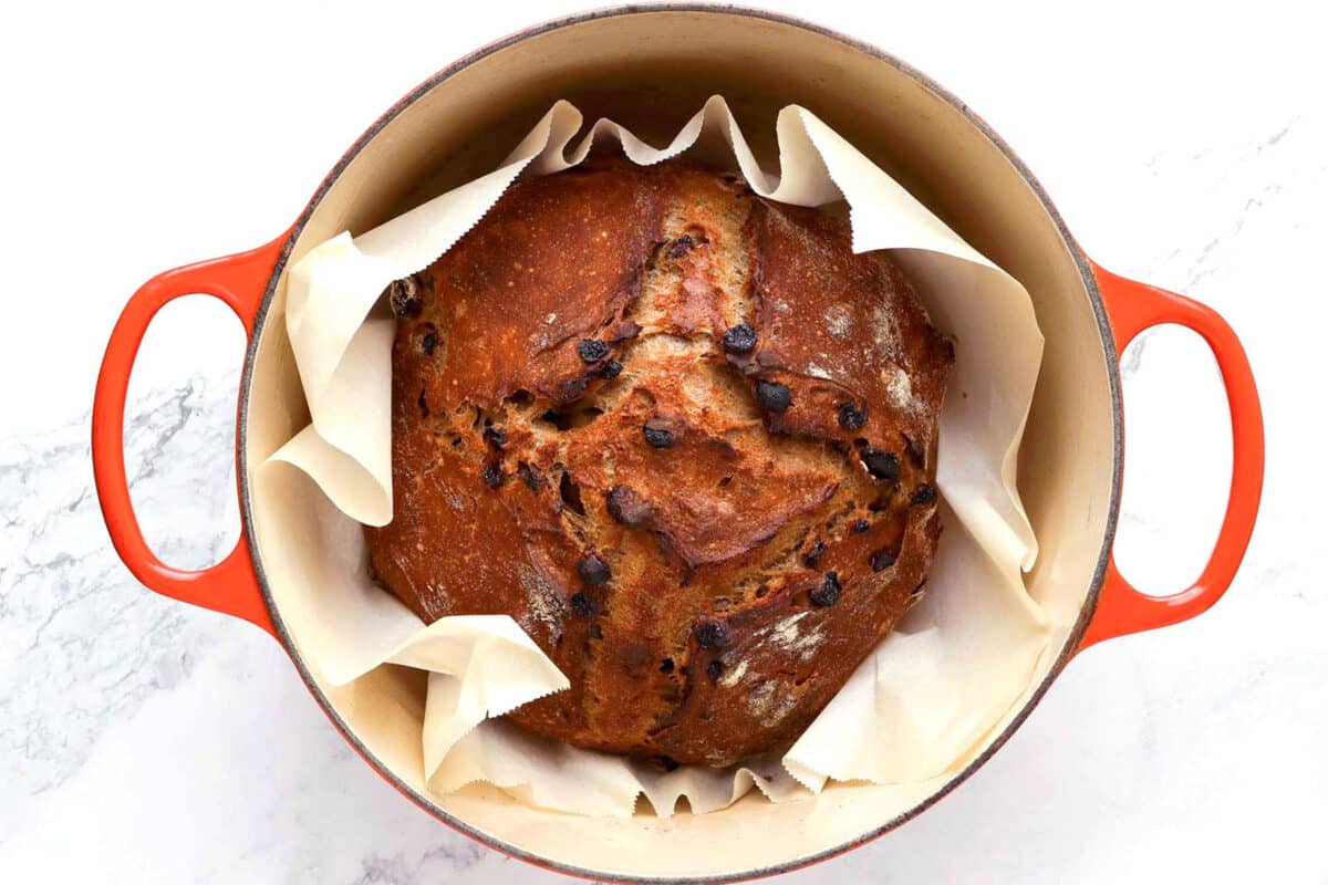 A round loaf of chocolate chip bread sits in a red Dutch oven lined with parchment paper, viewed from above on a white surface, reminiscent of a classic cinnamon raisin bread recipe with its inviting, home-baked charm.