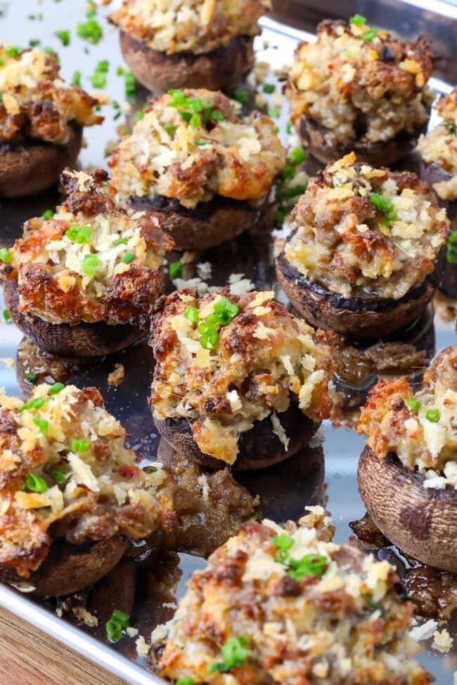 A close-up of spicy sausage stuffed mushrooms topped with golden breadcrumbs and chopped green herbs on a metal baking tray. The mushrooms are filled with a savory, crumbly mixture for the perfect sausage stuffed mushrooms recipe.