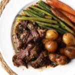 A white plate with shredded pot roast, baby potatoes, green beans, and glazed carrots sits on a woven placemat. The savory pot roast and tender veggies are all covered with a light sauce for a comforting meal.