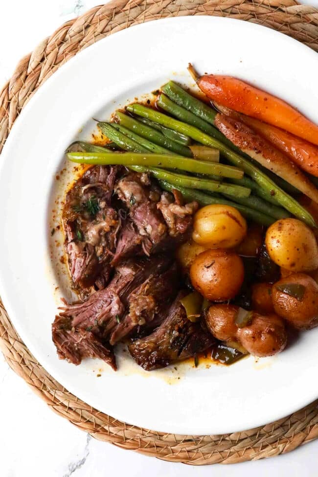 A white plate with shredded pot roast, baby potatoes, green beans, and glazed carrots sits on a woven placemat. The savory pot roast and tender veggies are all covered with a light sauce for a comforting meal.