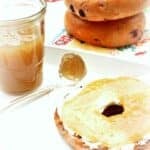 A toasted bagel half with cream cheese sits on a plate next to a spoon and a jar of pear honey. In the background, two whole bagels are stacked on a colorful napkin.