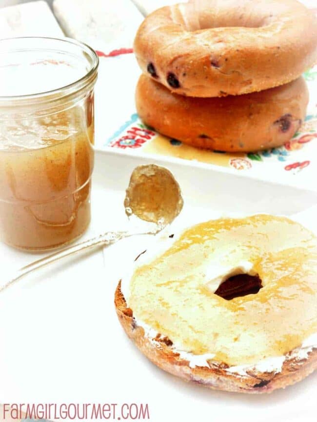 A toasted bagel half with cream cheese sits on a plate next to a spoon and a jar of pear honey. In the background, two whole bagels are stacked on a colorful napkin.