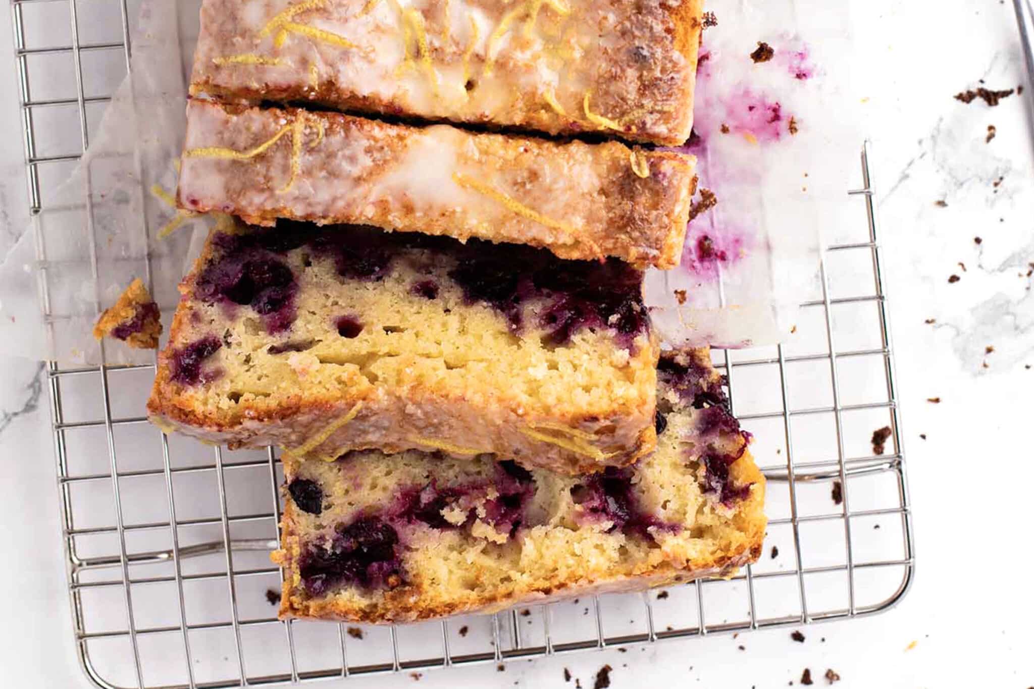 Sliced blueberry lemon loaf with icing on top, cooling on a wire rack. Blueberry pieces are visible in the moist yellow cake, with crumbs and icing scattered around.