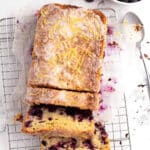 A blueberry lemon loaf with icing and lemon zest sits partially sliced on a wire rack. Nearby, a bowl of fresh blueberries and a spoon rest on the white surface, highlighting this classic lemon loaf treat.