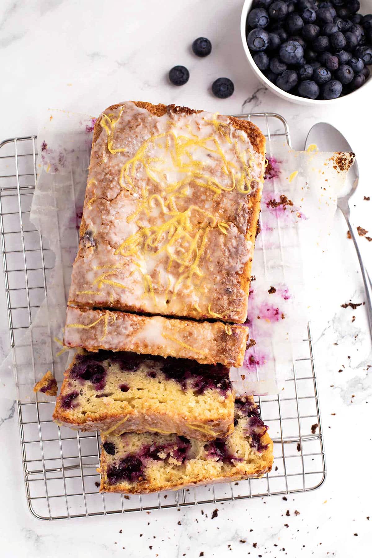 A blueberry lemon loaf with icing and lemon zest sits partially sliced on a wire rack. Nearby, a bowl of fresh blueberries and a spoon rest on the white surface, highlighting this classic lemon loaf treat.