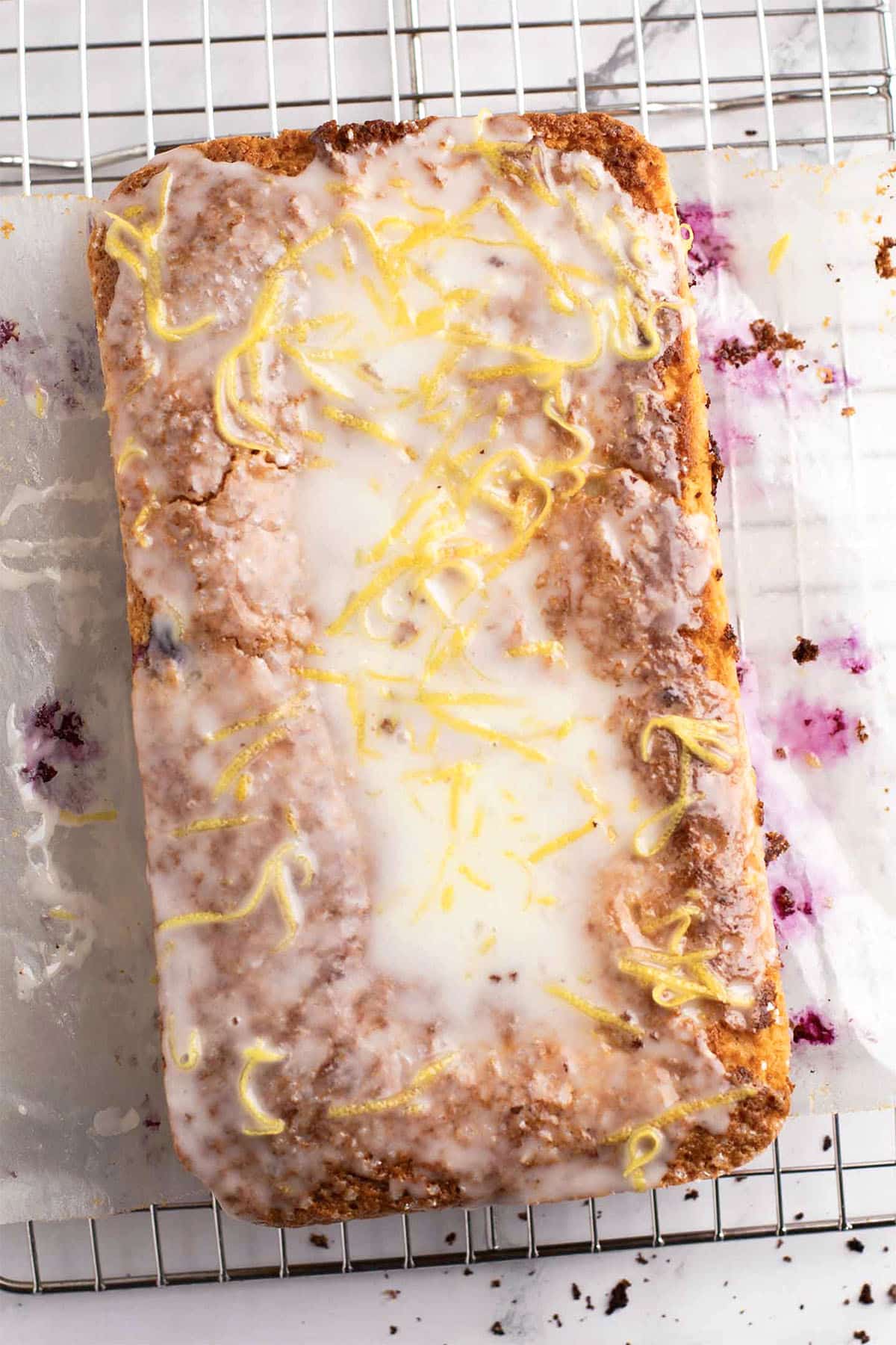 A lemon loaf, glazed and topped with fresh lemon zest, sits on a wire cooling rack with crumbs and icing drips scattered on the parchment paper below.