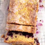 A blueberry lemon loaf with glaze and lemon zest on top, partially sliced on a wire cooling rack lined with parchment paper.