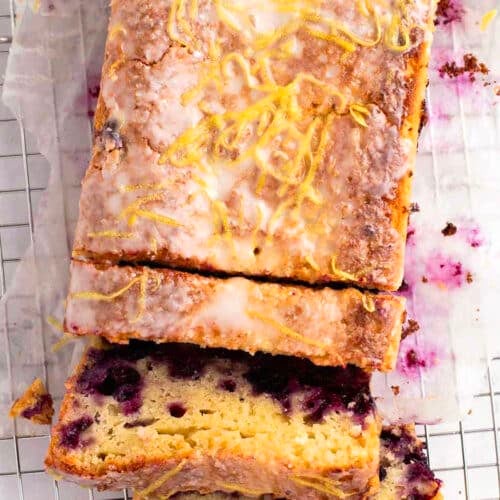 A blueberry lemon loaf with glaze and lemon zest on top, partially sliced on a wire cooling rack lined with parchment paper.