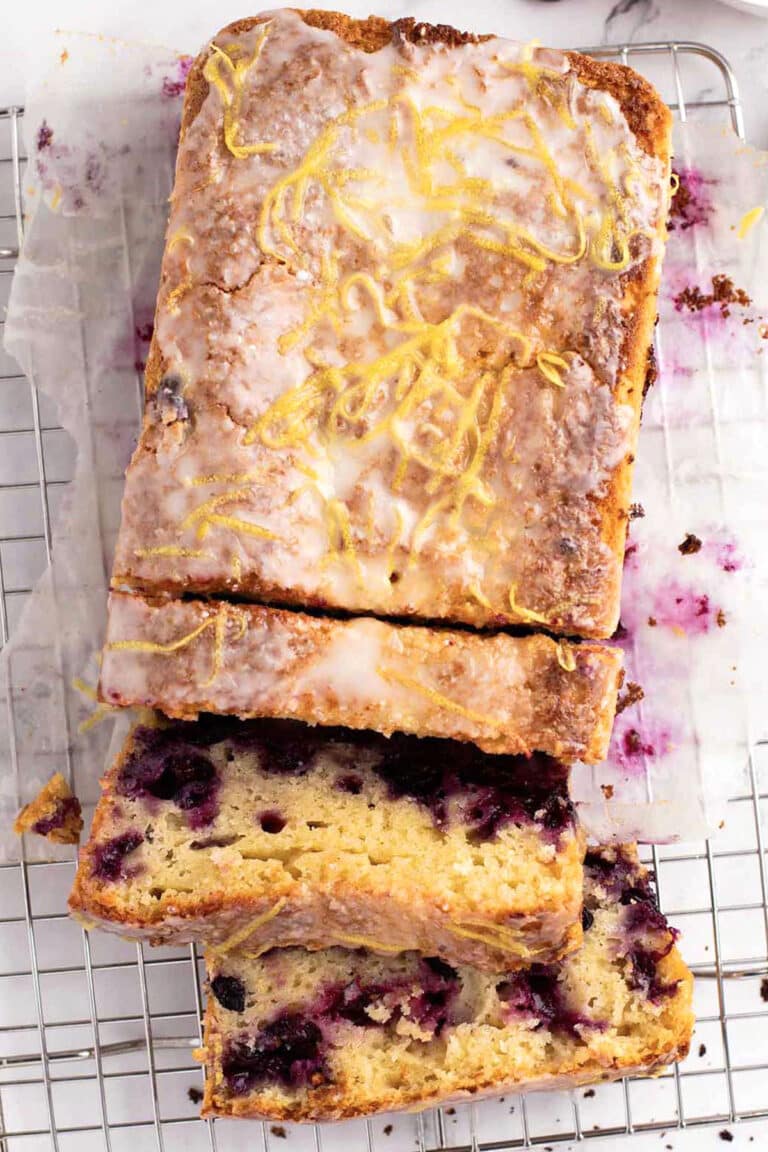 A blueberry lemon loaf with glaze and lemon zest on top, partially sliced on a wire cooling rack lined with parchment paper.