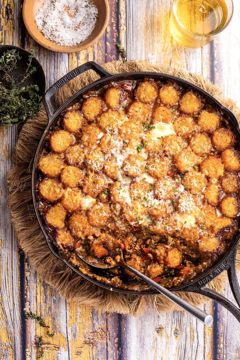 A cast iron skillet filled with a hearty tater tot casserole, topped with melted cheese, is shown on a rustic wooden surface with a serving spoon, fresh herbs, salt, and a glass of white wine nearby.