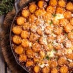Top-down view of a cast-iron skillet filled with a golden tater tot casserole, topped with melted cheese, sprinkled herbs, and parmesan. Beside it are fresh thyme sprigs, resting on a textured woven mat against a rustic wooden background.