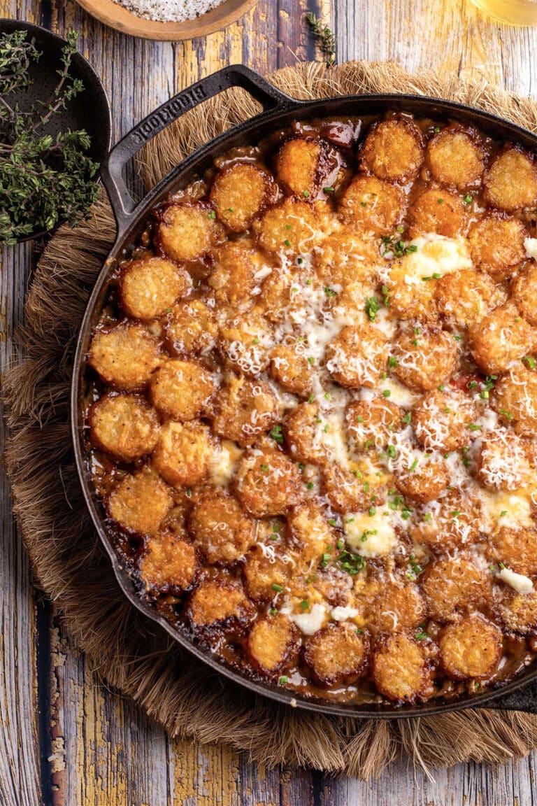 Top-down view of a cast-iron skillet filled with a golden tater tot casserole, topped with melted cheese, sprinkled herbs, and parmesan. Beside it are fresh thyme sprigs, resting on a textured woven mat against a rustic wooden background.