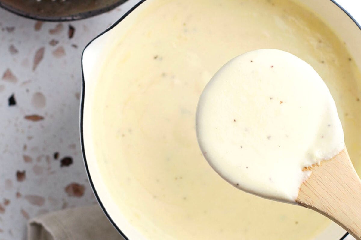 A wooden spoon holds up a thick, creamy white homemade alfredo sauce with black pepper specks over a white saucepan filled with more of the same sauce, resting on a speckled countertop. This is alfredo sauce that doesn't break.