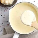 A saucepan filled with creamy homemade alfredo sits on a beige cloth, with a wooden spoon lifting some sauce. In the background, a bowl of grated cheese and a wedge of cheese complete this easy alfredo sauce recipe scene.