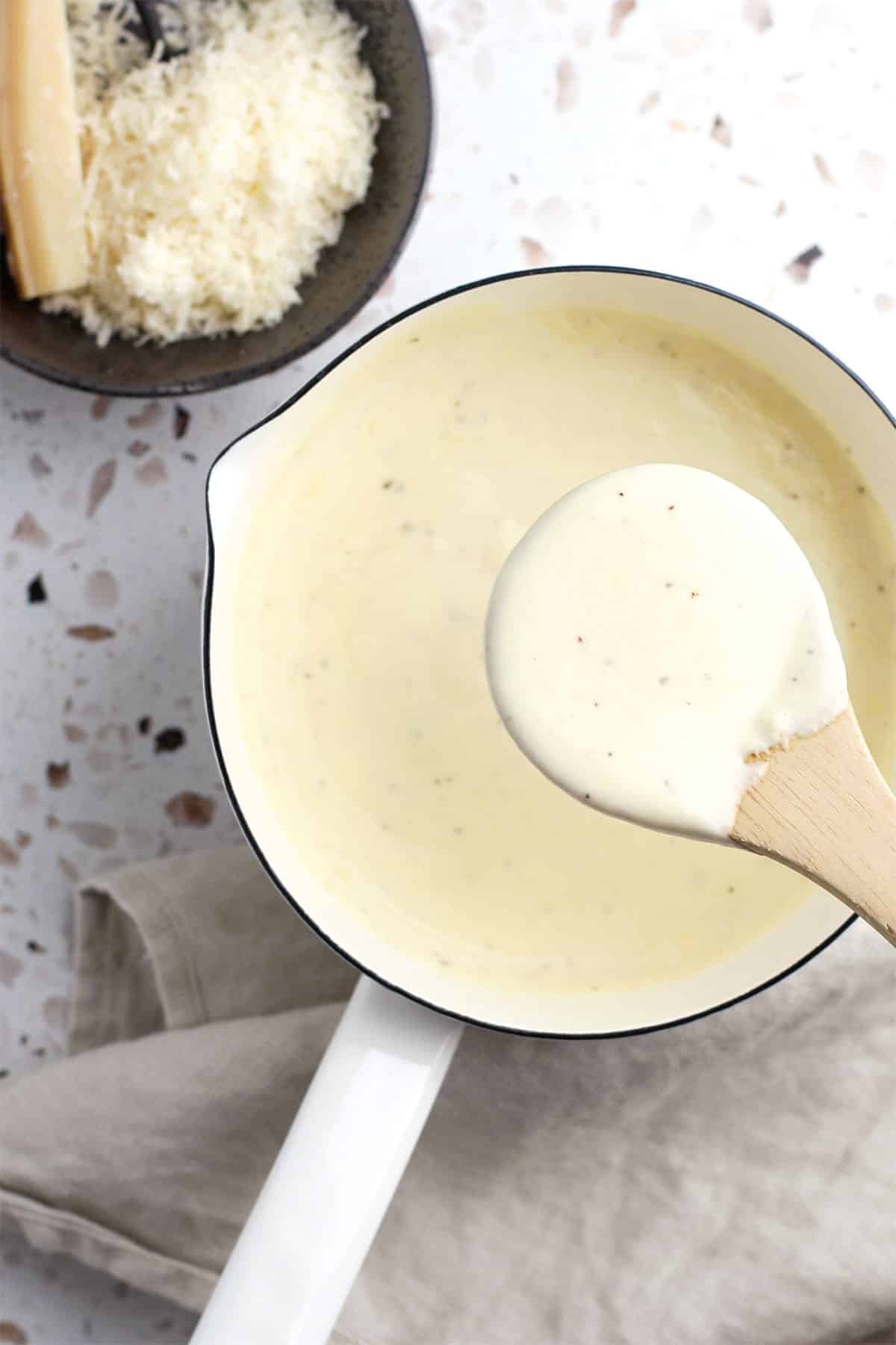 A saucepan filled with creamy homemade alfredo sits on a beige cloth, with a wooden spoon lifting some sauce. In the background, a bowl of grated cheese and a wedge of cheese complete this easy alfredo sauce recipe scene.