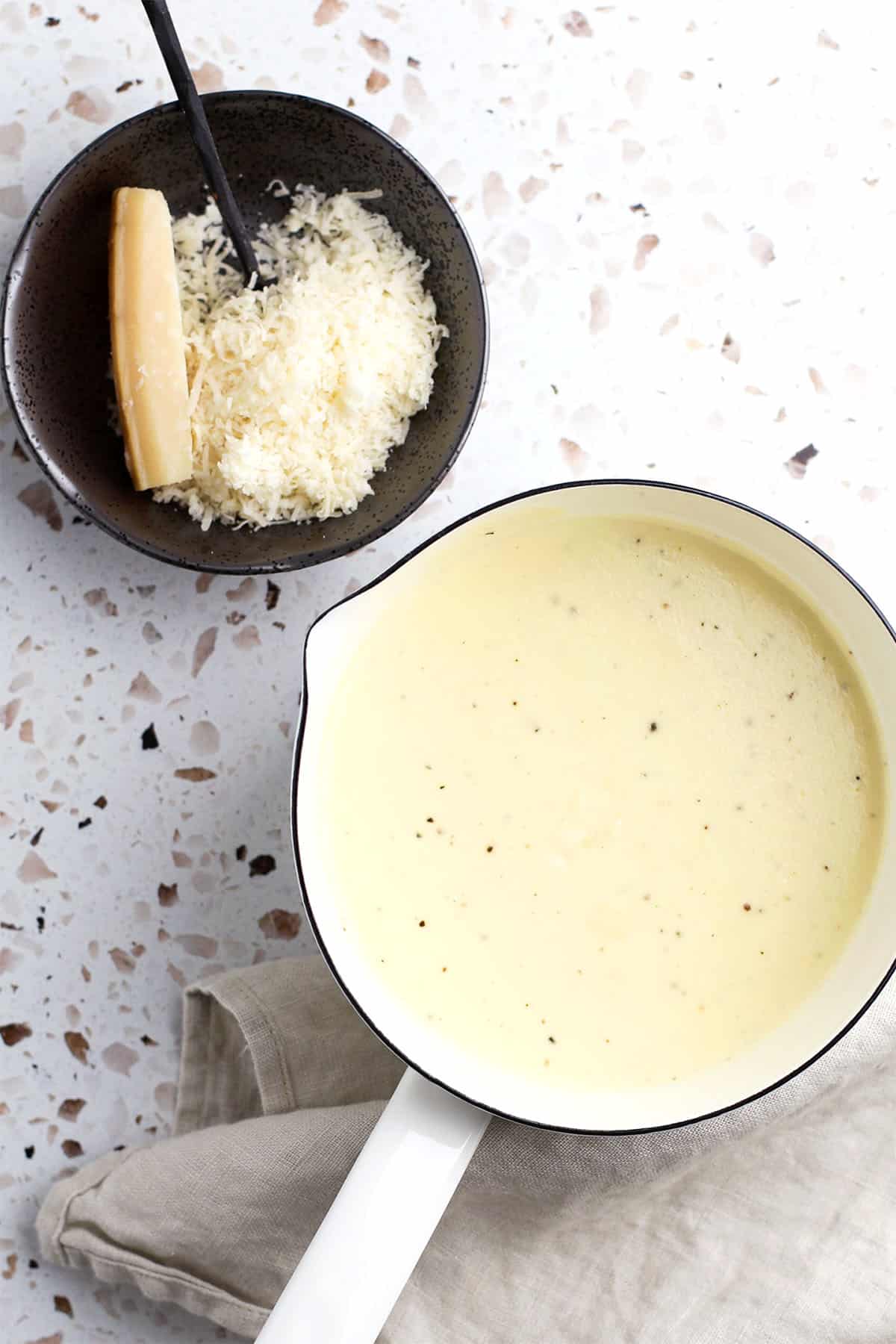 A white saucepan filled with creamy homemade alfredo sauce sits on a light surface beside a bowl of grated cheese and a wedge of Parmesan cheese, with a spoon resting in the bowl.